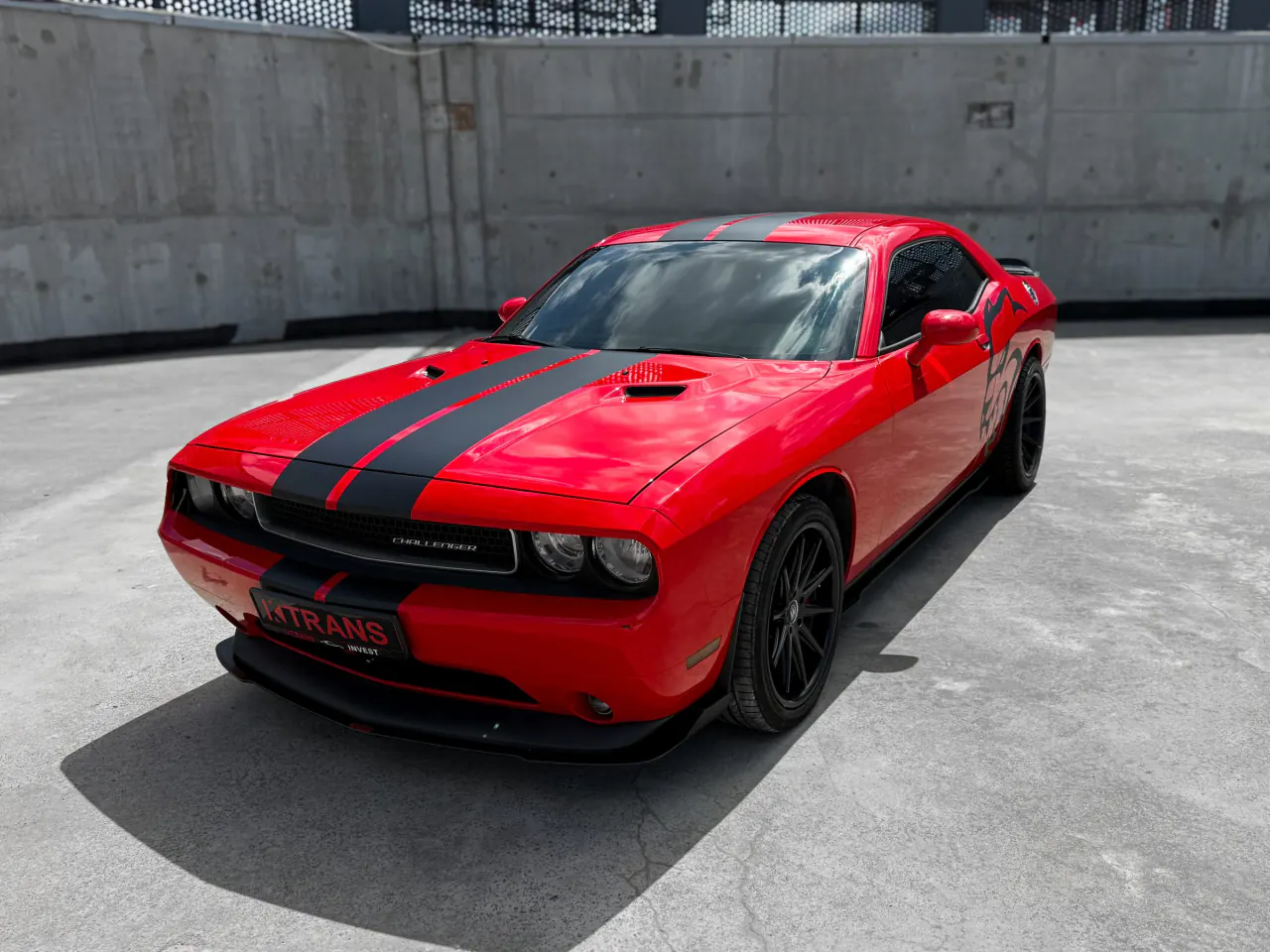 Красный Dodge Challenger 2013 с черными полосами и табличкой KTRANS на бетонной крыше паркинга